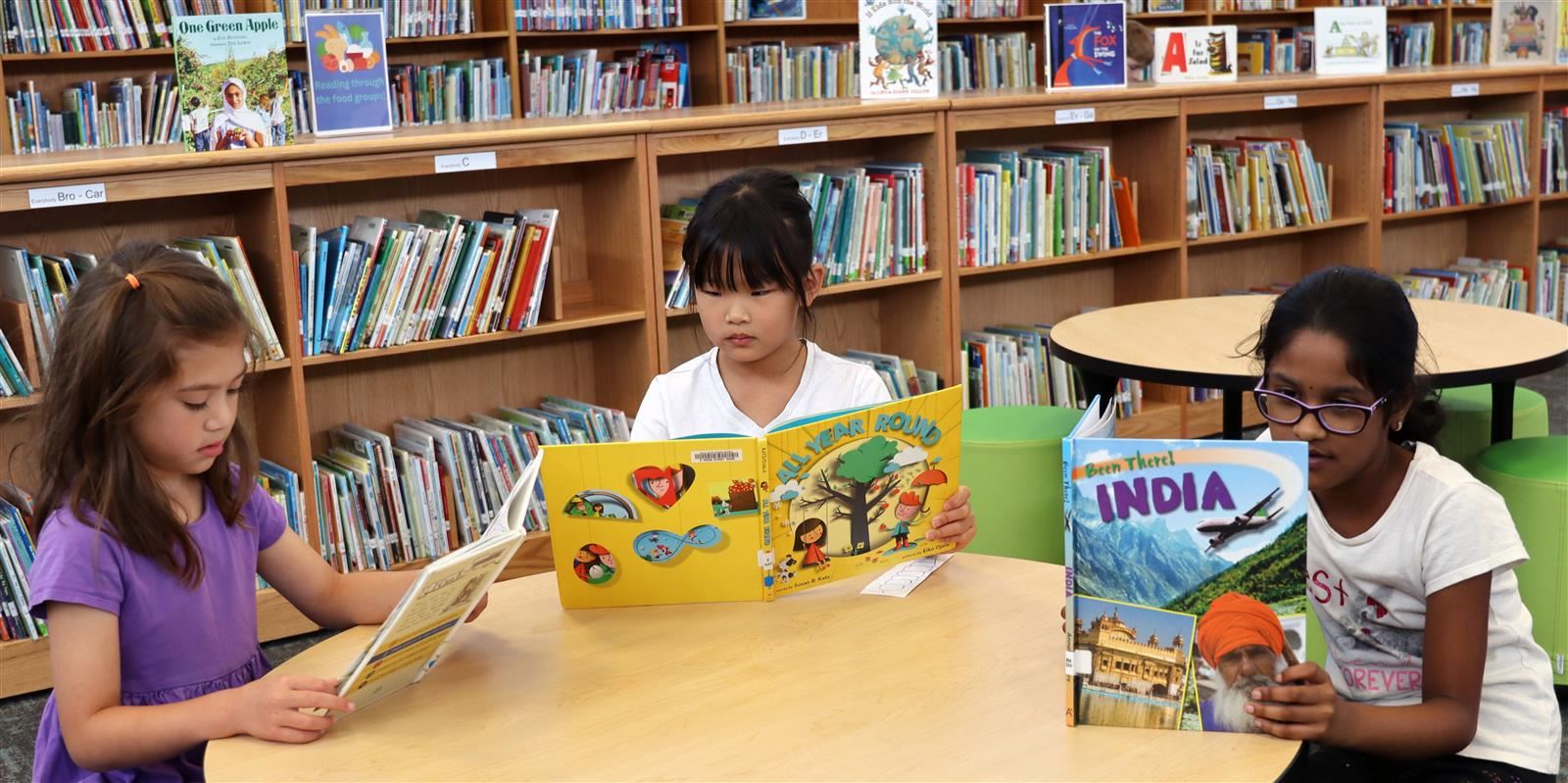 Three elementary students sit at a round table reading books in a library.